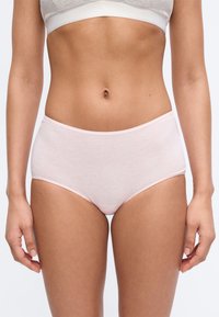 Γυναίκα που φοράει ανοιχτό ροζ βαμβακερά σλιπ ψηλόμεσα και λευκό αθλητικό σουτιέν, στέκεται μπροστά σε απλό ουδέτερο φόντο.