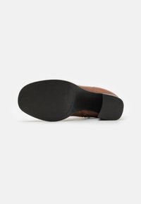 Zapato de piel marrón con suela de goma, que presenta un patrón texturizado y un tacón bajo, mostrando un detalle de costura a lo largo del costado.
