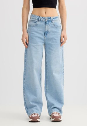 Persona vistiendo vaqueros anchos de pierna en azul claro, top corto negro y zapatillas rosa y blancas, de pie sobre fondo blanco.