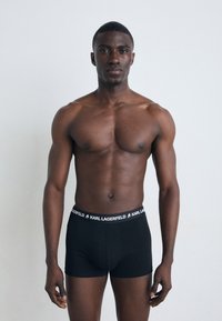 Boxers negros ajustados con una cinturilla elástica de marca, usados por un modelo masculino de complexión musculosa, sobre un fondo neutro.