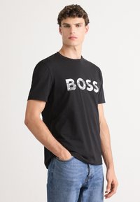 Schwarzes Baumwoll-T-Shirt mit kurzen Ärmeln, das einen großen, gittermusterartigen "BOSS"-Text in Weiß zeigt. Kombiniert mit hellblauen Jeans.