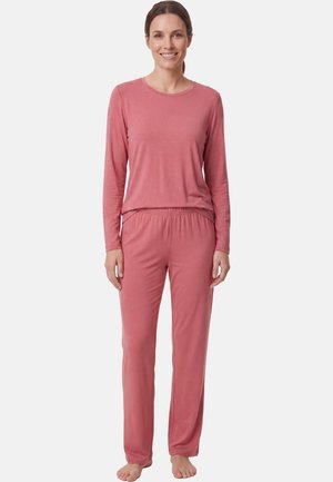 Lachende vrouw staat blootsvoets en draagt een bijpassende zachte roze longsleeve en losse broek tegen een effen lichte achtergrond.