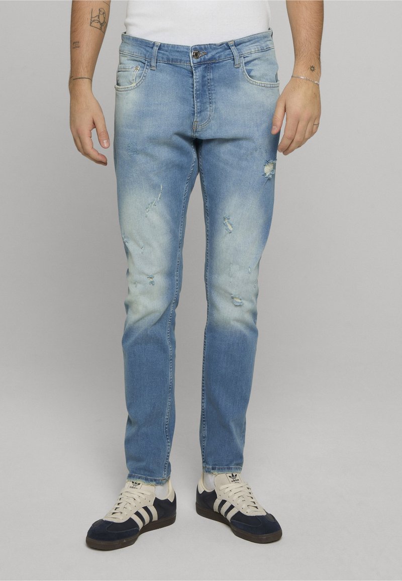 Světle modré džíny s úzkým střihem, zdobené efektu vyšisování a opraných míst. Nošeny s černo-bílými teniskami.