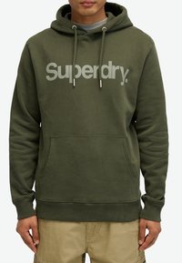 Mørk grøn hættetrøje lavet af bomuld med en frontlomme, snoretræk i hætten og et lysegråt "Superdry" logo. Ribstrikkede manchetter og søm.
