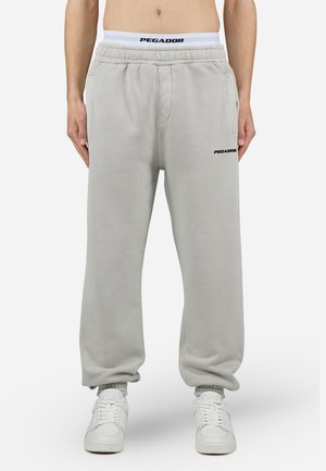 Pantaloni della tuta grigi chiari con cintura elastica, tasche laterali e logo "PEGADOR" nero. Gambe a sigaretta con polsini elastici. Tessuto morbido.
