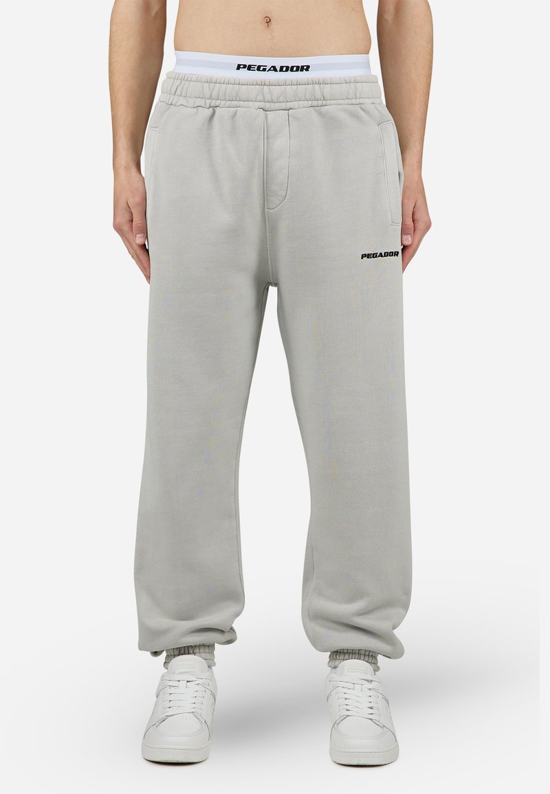 Pantalones de chándal gris claro con cinturilla elástica, bolsillos laterales y un logotipo negro "PEGADOR". Piernas ajustadas con puños elásticos. Textura de tela suave.