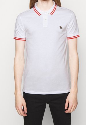 Person iført en hvid polo-shirt med røde striber på kraven og ærmekanterne, lille broderet zebra-logo på venstre bryst, sorte bukser.