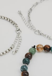 Trois bracelets : une chaîne en argent avec des maillons ronds, une chaîne en argent avec un fermoir à homard, et un bracelet en perles avec des pierres marron, vertes et bleues.