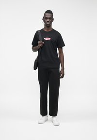 Melna kokvilnas T-krekls ar "Guess Jeans" logotipu, kombinēts ar melnām šaurām biksēm un baltām sporta kurpēm. Modelis stāv ar melnu plecu somu.