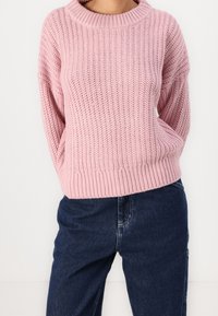 Jasnoróżowy dzianinowy sweter z szerokim, żeberkowym dekoltem i długimi rękawami, o luźnym kroju i szczegółach teksturalnych. W zestawie z ciemnoniebieskimi dżinsami.