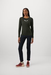 Long-sleeve olive green top with a white logo, paired with navy pants and red sneakers. The fabric appears smooth and fitted, with a crew neckline.