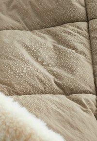 Manta beige acolchada con gotas de agua en la superficie, presenta un suave borde de piel blanca a lo largo de un lado, destacando la textura y el contraste.