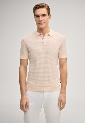 Giovane uomo che indossa una maglietta polo a maniche corte in tessuto leggero color pesca chiaro e pantaloni bianchi, in piedi davanti a uno sfondo chiaro e uniforme.