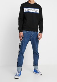 Czarna bluza z kapturem z biało-niebieskim, paskowanym logo, zestawiona z jasnoniebieskimi dżinsami, z brandingiem po bokach i niebieskimi trampkami.