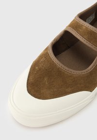 Pantof maro din piele întoarsă cu talpă albă de cauciuc, cu vârf rotund și două secțiuni decupate pe partea superioară pentru un design unic.