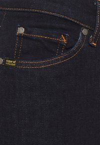 Mörkblå denimjeans med kontrasterande orange sömmar. Har en framficka, metalldetaljer och en liten svart etikett märkt "Tiger of Sweden."