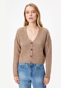 Cardigan marrón con canalé, con un escote en V profundo, botones en la parte delantera y mangas largas. Se lleva con jeans de cintura alta de color azul claro. Textura suave.