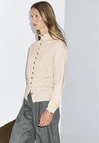 Cardigan en maille beige clair avec col haut, boutons noirs contrastants et manches longues ; associé à un pantalon rayé gris.