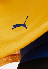 Žlutá látka s modrým logem Puma, s hladkým povrchem. Vrstevnatý design ukazuje tmavě modré a černé materiály pod ním.