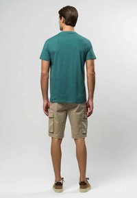 Zielony tekstylny T-shirt z krótkimi rękawami, zestawiony z beżowymi szortami typu cargo z bocznymi kieszeniami, noszony z brązowymi butami na płaskiej podeszwie.