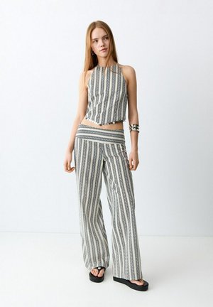 Mladi žena nosi odgovarajući crno-bijeli top na bretele s vertikalnim prugama i široke hlače, stoji na bijeloj podlozi uz običan zid.