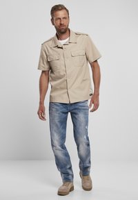 Camicia beige a maniche corte con bottone davanti, due tasche sul petto; abbinata a jeans chiari strappati e stivali beige.