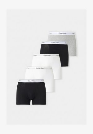 Slanke donkerhuidige man staat blootsvoets, draagt witte Calvin Klein boxershorts en kijkt lichtjes opzij tegen een effen lichte achtergrond.