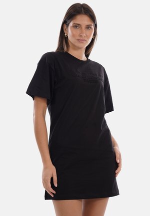 Mujer con cabello castaño liso que lleva pendientes dorados y un vestido negro suelto de manga corta con texto sutil en relieve en el pecho.