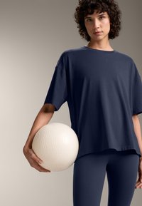 Tricou din bumbac oversized de culoare bleumarin, cu o textură fină, asortat cu colanți bleumarin. Ținând în mână o minge albă, texturată, cu modele ondulate.