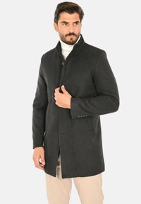 Cappotto di lana grigio scuro con collo alto, chiusura con un solo bottone, tasche laterali e una trama liscia, indossato sopra un dolcevita chiaro.