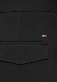 Zwarte stof met een zakdetail, voorzien van een kleine gouden hardwareaccent met rode en witte componenten. Gladde textuur, op maat gemaakte ontwerp.