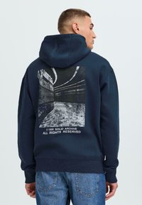 Námořnická modrá mikina s kapucí z měkkého materiálu, na zadní straně s grafickým potiskem v černobílém abstraktním designu a bílým textem.
