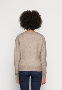 Osoba o kręconych włosach, ubrana w beżowy sweter z dzianiny i ciemnoniebieskie dżinsy, pokazana od tyłu na jednolitym tle.