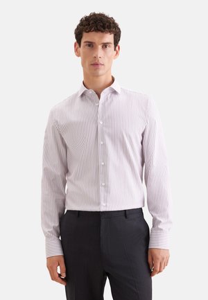 Homme portant une chemise boutonnée à rayures verticales blanches et rose clair, et un pantalon noir, debout devant un fond uni.