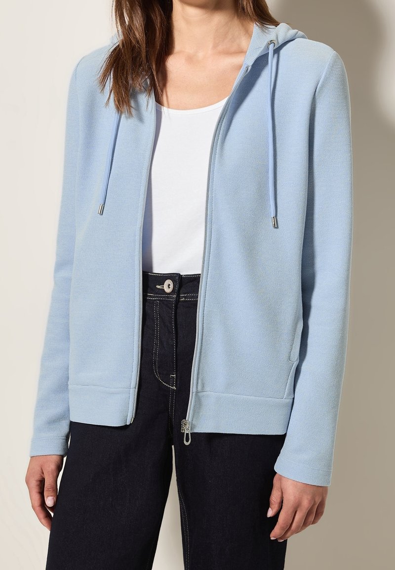 Femme portant un sweat à capuche bleu clair zippé sur un haut blanc et un pantalon foncé, debout devant un fond uni.