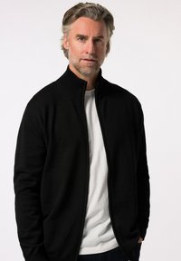 Cardigan negro con cremallera, hecho de material de punto suave, con un cuello alto y mangas largas, llevado sobre una camiseta blanca. Diseño sencillo.
