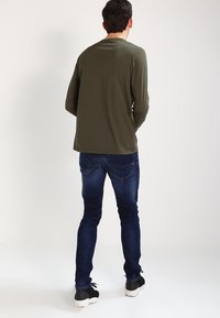 Olivgrön långärmad tröja med rund halsringning, i kombination med mörkblå slim-fit jeans och svarta träningsskor, sedd bakifrån.