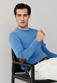 Młody mężczyzna siedzący na czarnym krześle, ubrany w niebieski sweter z brązowymi łatami na łokciach i białe spodnie, patrzący w kamerę.