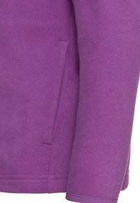 Manche de veste polaire violette et poche latérale avec couture visible et tissu à texture douce.