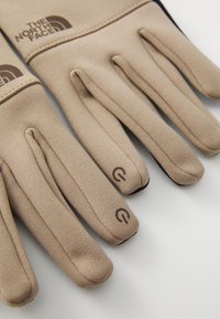 Beige hansker laget av elastisk stoff med en glatt tekstur. De har en brun logo og detaljer på mansjettene og fingertuppene.