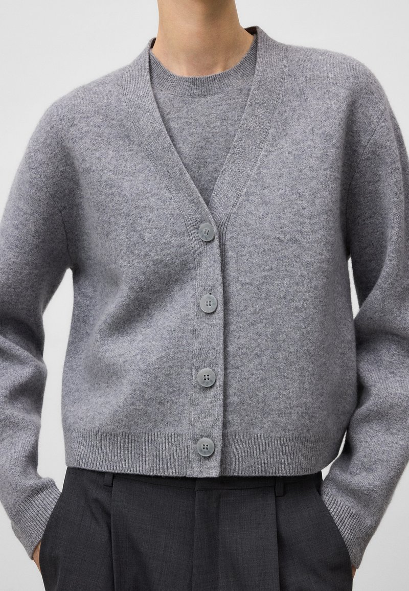 Grijze wollen cardigan met een V-hals en vijf knopen, met lange mouwen en een verkorte zoom, gelaagd over een bijpassende grijze trui.