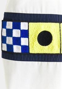 Tela blanca con un parche horizontal que presenta un diseño de cuadros en azul y blanco, un cuadrado amarillo con un círculo negro, bordeado en azul marino. La textura es variada.