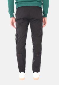 Pantaloni cargo neri in cotone con tasche laterali e dettagli in bottone, abbinati a sneakers bianche. Vista posteriore che mostra vestibilità e design.