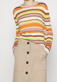 Pull en tricot avec des rayures horizontales orange, rose, jaune et marron ; il a un col rond. Assorti à une jupe beige avec des boutons noirs.