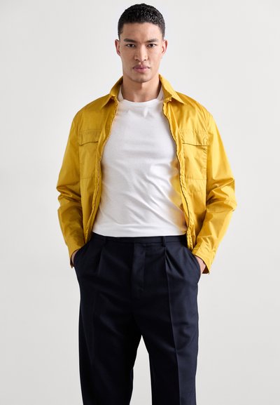 Veste légère jaune avec un col, deux poches poitrine, portée sur un t-shirt blanc, associée à un pantalon noir ajusté.