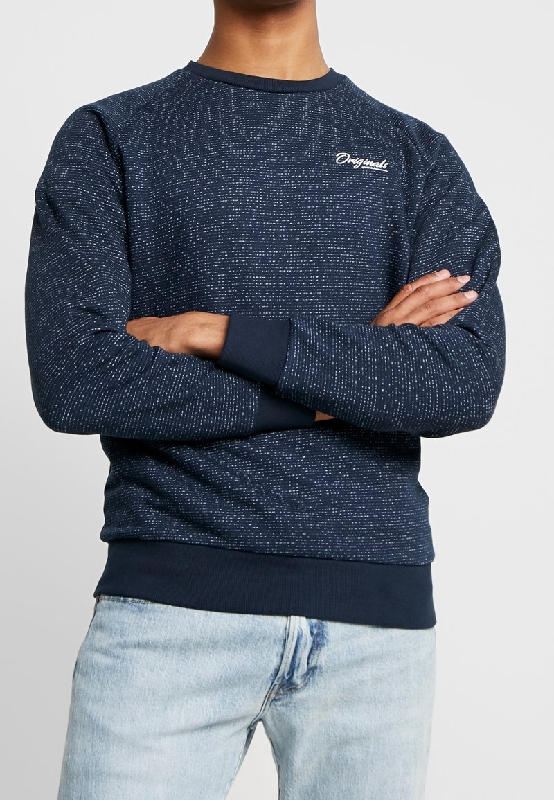Homme bras croisés portant un sweat-shirt bleu marine moucheté avec le texte "Originals" et un jean bleu clair.