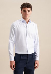 Camicia da uomo azzurro chiaro a maniche lunghe con colletto a punta, realizzata in tessuto morbido. Indossata con pantaloni blu navy su misura.