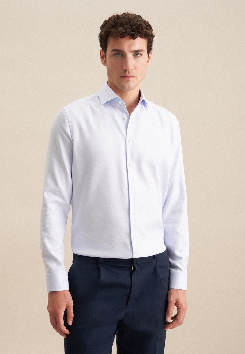 Camicia da uomo azzurro chiaro a maniche lunghe con colletto a punta, realizzata in tessuto morbido. Indossata con pantaloni blu navy su misura.