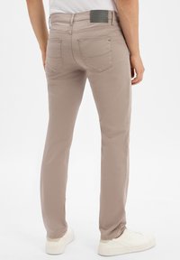 Mężczyzna w beżowych dżinsach o prostym kroju i białych trampkach, stojący z lekko rozstawionymi nogami na jednolitym białym tle.