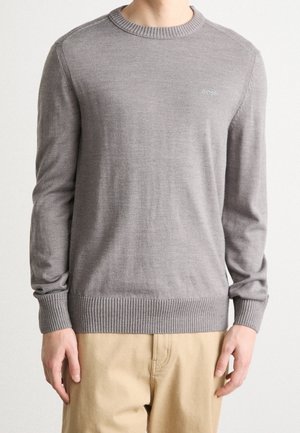 Pull gris à manches longues avec col rond, poignets et ourlet côtelés, porté par une personne en pantalon beige, avec un petit logo "BOSS" sur la poitrine.